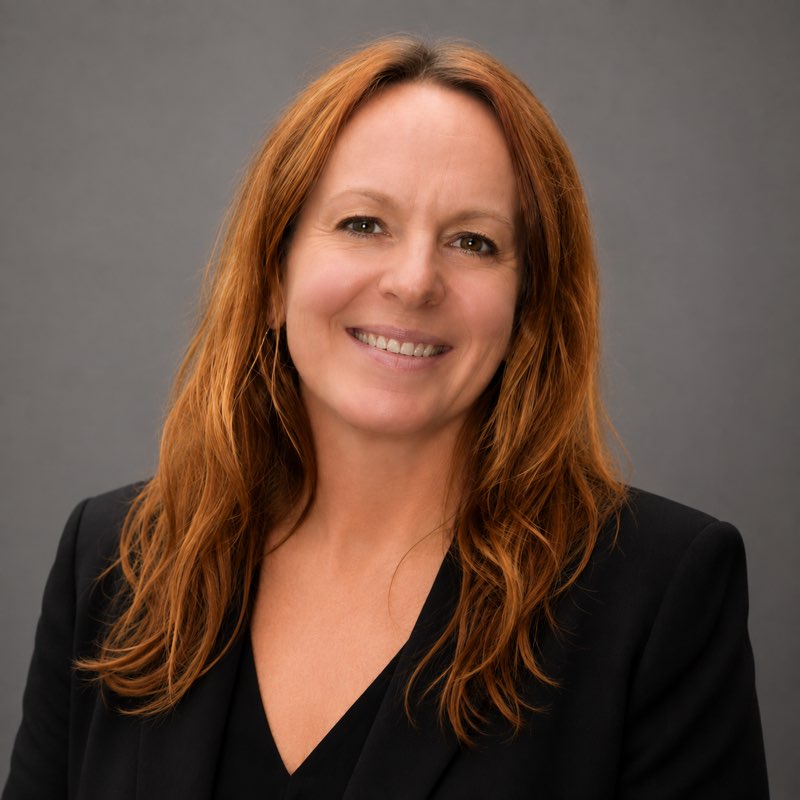 Smiling woman with red hair wearing a black blazer against a gray studio backdrop (professional headshot).