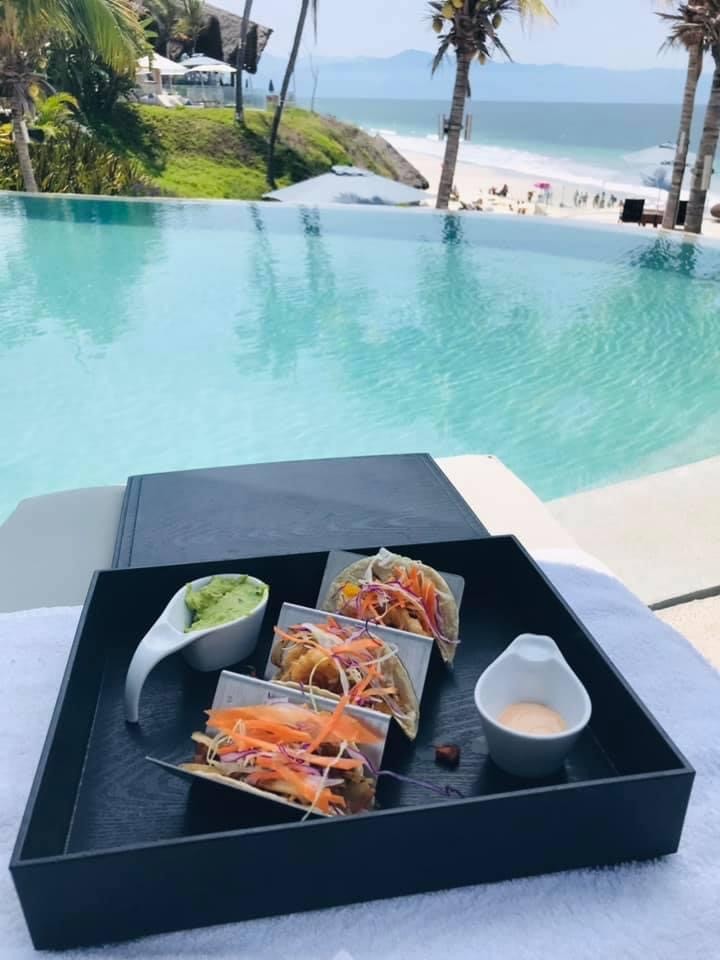Three tacos with colorful toppings on a black tray beside an infinity pool, beach and palm trees in the background.