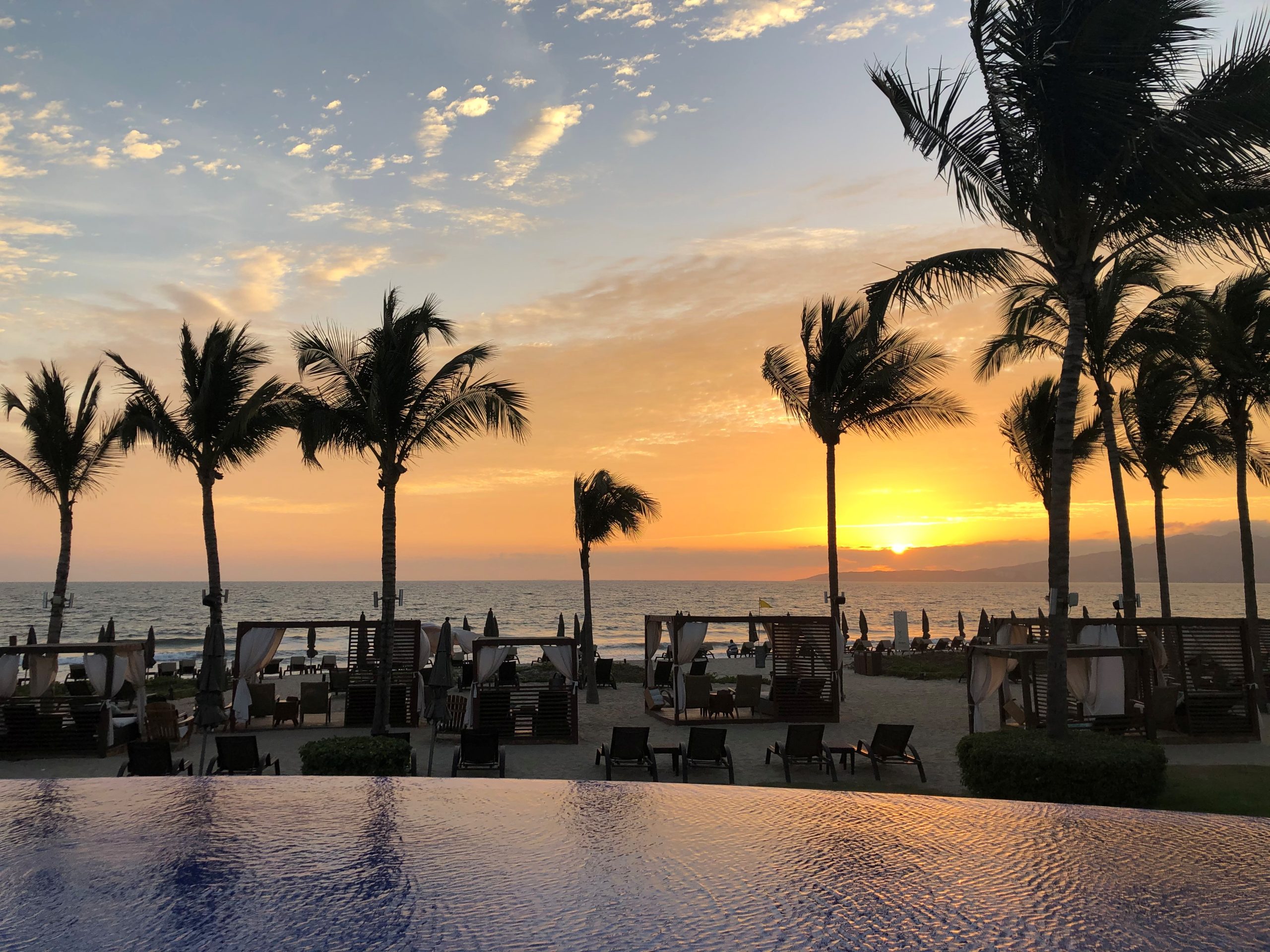 Sunset over a beach with silhouetted palm trees, lounge chairs and umbrellas along the shore reflecting in the calm water. The foreground shows a pool and cabanas.