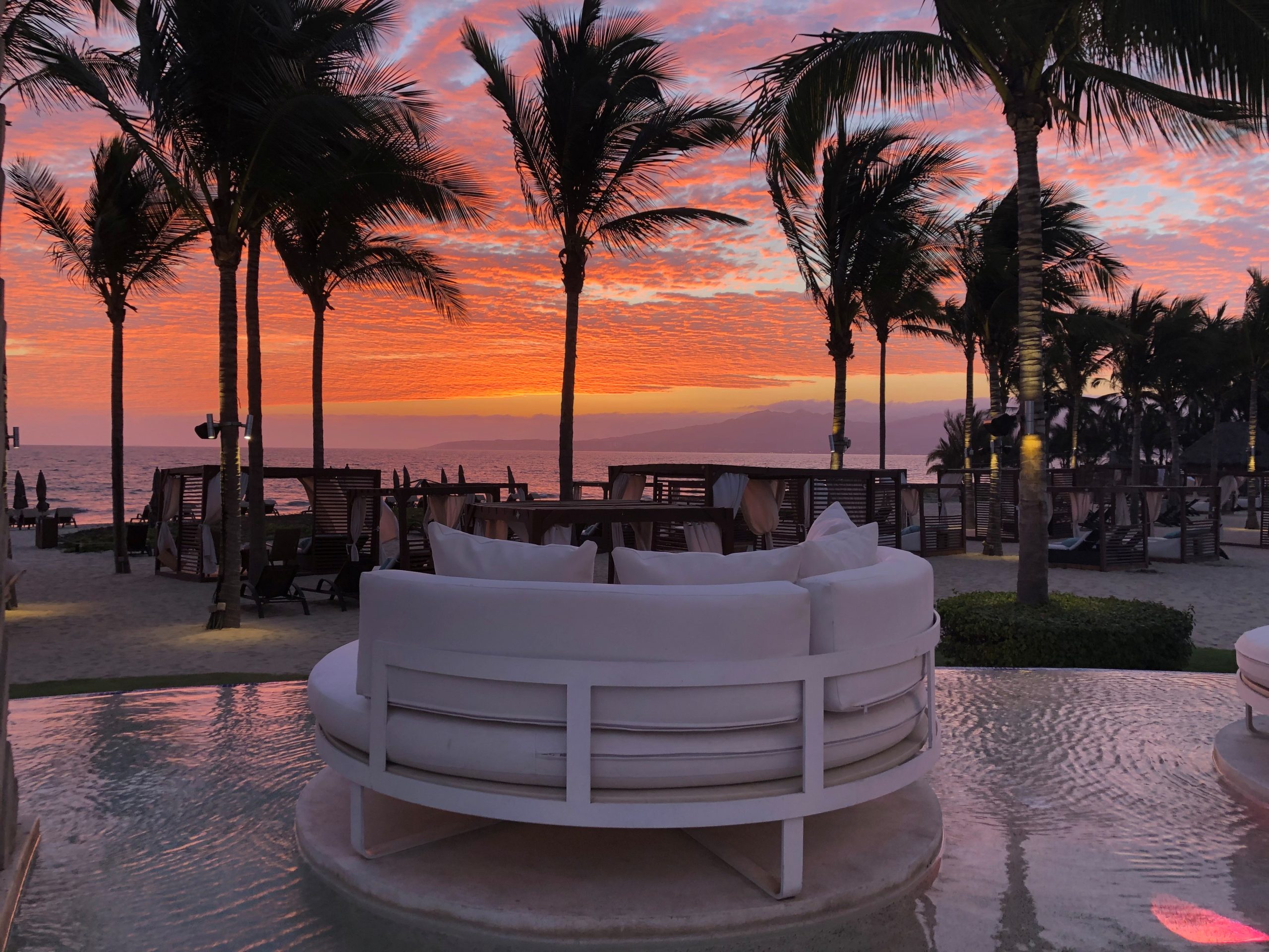 Sunset over a tropical beach with silhouetted palm trees and white circular lounge seating by the water.