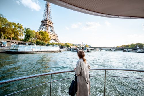 Person in a beige coat standing at a railing on a Seine riverboat, gazing toward the Eiffel Tower in Paris.