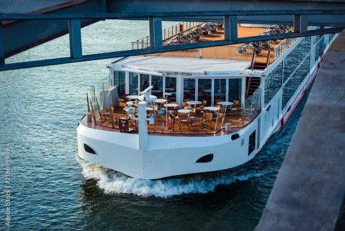 White cruise boat cruising under a bridge, with outdoor dining on the upper deck and lounge chairs visible on the sun deck.