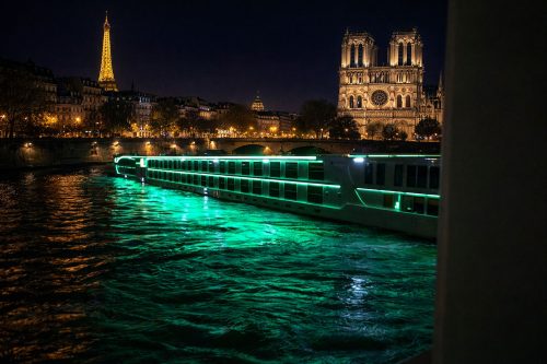 Nighttime Paris scene: Notre-Dame Cathedral on the right, the Eiffel Tower glowing in the distance, and a green-lit boat on the Seine.