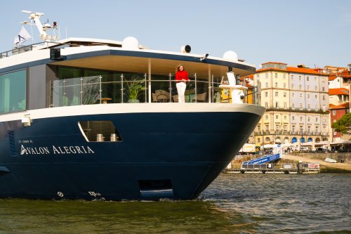 Bow of a luxury yacht named Avalon Alegria, with a woman in a red sweater standing on the upper deck as the harbor and colorful buildings loom behind.