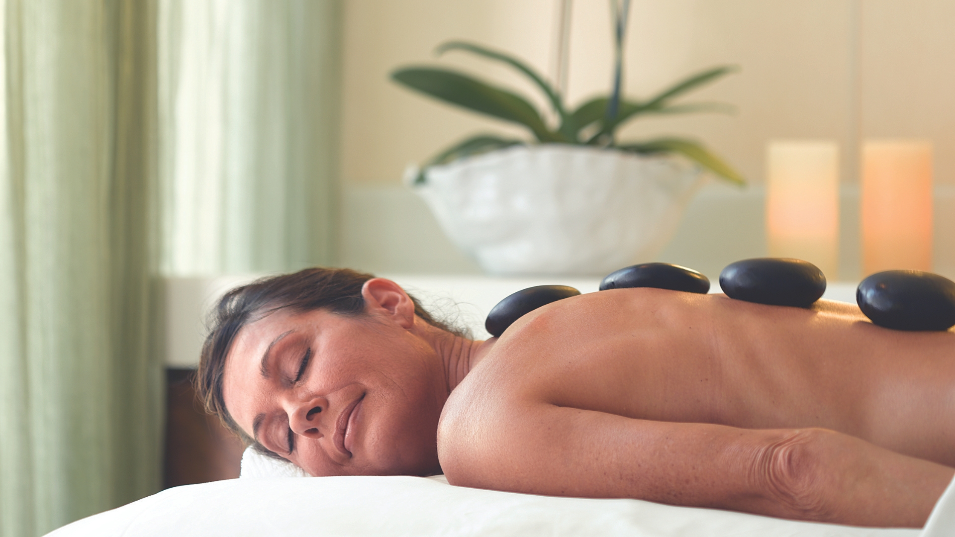 Woman lying face down, receiving a hot stone massage on her back in a spa setting.