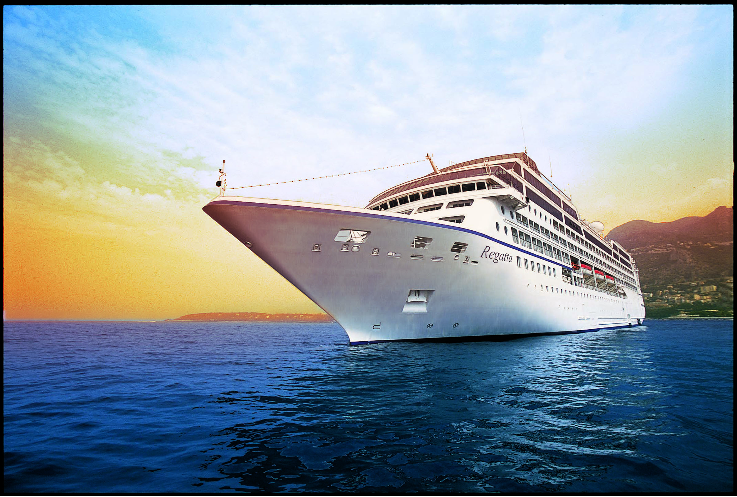 Large white cruise ship named Regatta docked or anchored on calm blue water during a colorful sunset with distant hills in the background