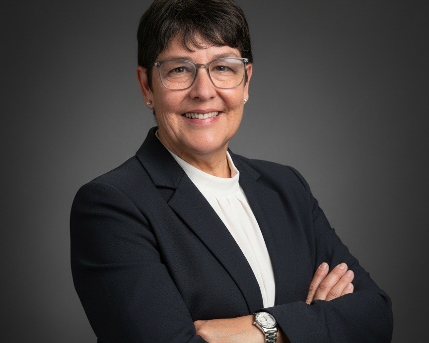 Professional headshot of a smiling woman in a dark blazer with arms crossed against a gray backdrop.