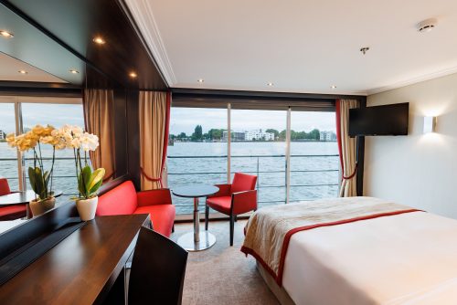 Hotel-like cruise cabin with red seating, a bed, a desk, and large windows overlooking water.