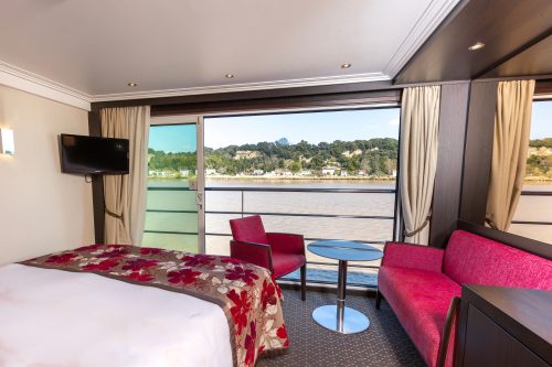 Hotel room with a bed in foreground, red seating, and a round blue table; large windows overlook a river and tree-covered hills outside.