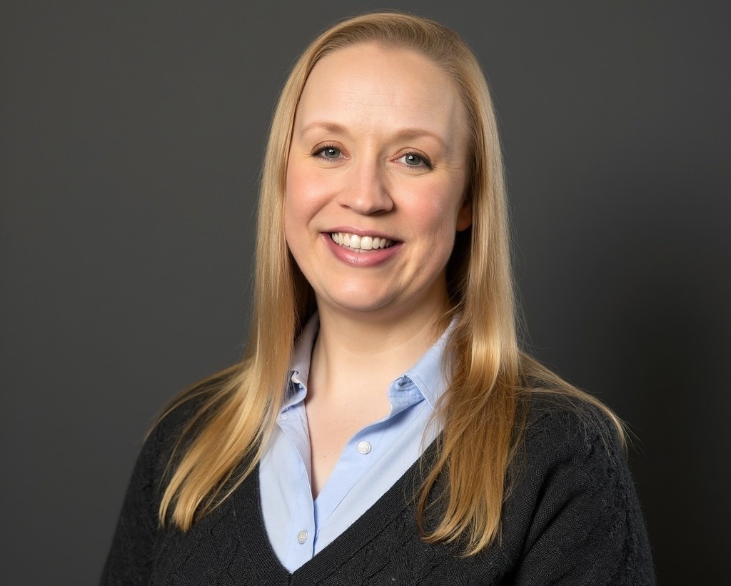 Rachel Mesich | Holiday Planit Smiling blonde woman in a light blue blouse and black sweater, posed for a professional headshot against a gray background.