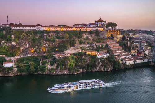 White multi-deck river cruise ship sailing along a green, rocky hillside with a historic fortress and domed building lit at sunset on the opposite riverbank