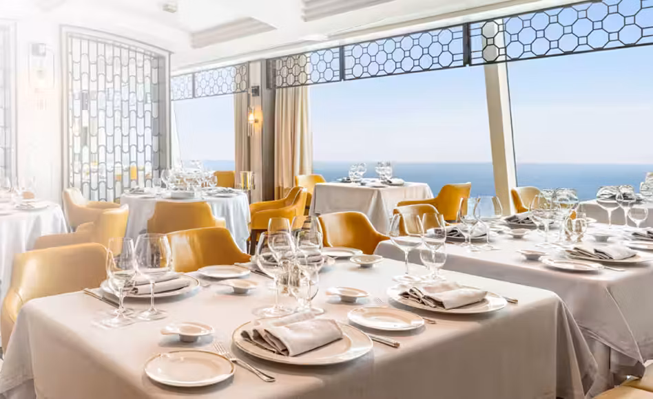 Elegant seaside restaurant with white-tablecloth tables set with plates, glassware, and yellow chairs, overlooking the ocean