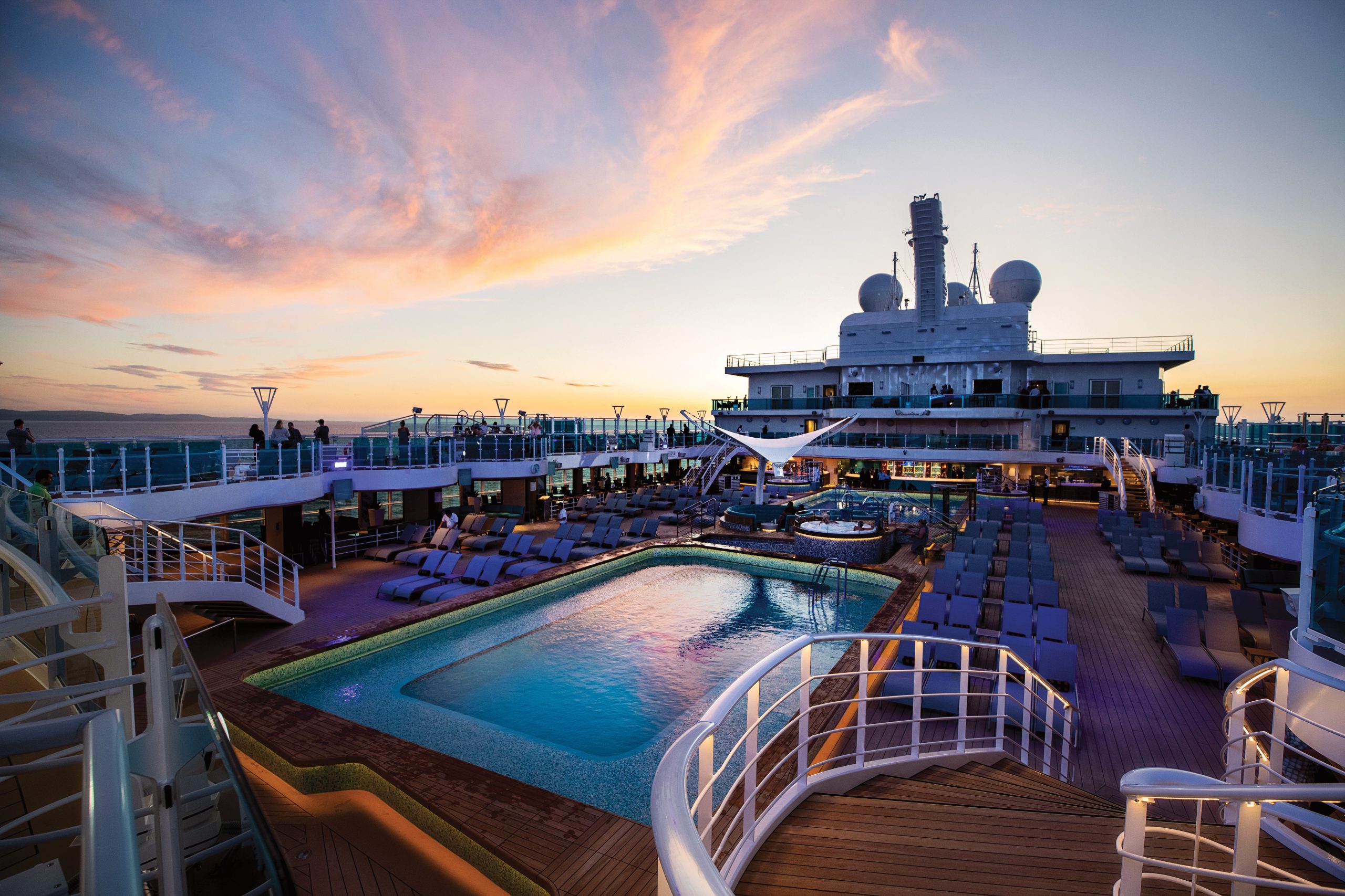Cruise ship pool deck at sunset with lounge chairs, glass railing, and a large ship structure in the background.