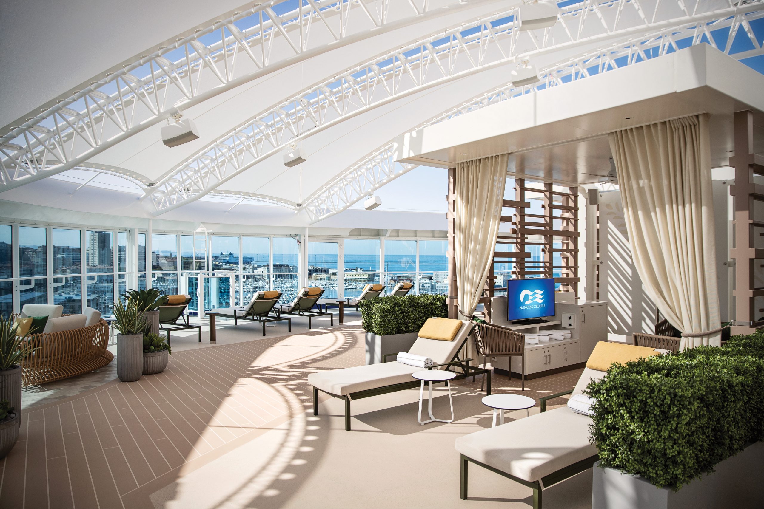 Open-air cruise lounge with white curved truss structures, beige curtains, and rows of lounge chairs overlooking the ocean and cityscape.