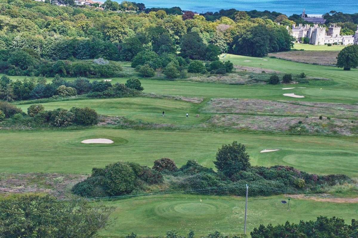 Priscila Gonzalez Hernandez | Holiday Planit Aerial view of a lush seaside golf course with green fairways, sand bunkers, trees, and a distant castle along the horizon by the blue sea.