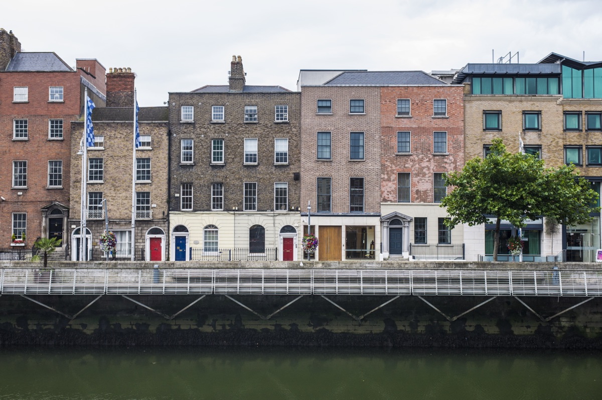 Historical center of Dublin Quay of the river liffe | Holiday Planit Row of brick townhouses along a riverfront with colorful doors and hanging plants, a pedestrian walkway, and a metal railing in front of the water.
