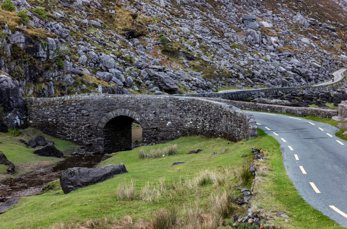 EU15 CHA0107 | Holiday Planit Stone arched bridge over a narrow stream with a winding road beside a rocky hillside.